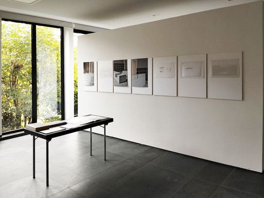 Foto eines Ateliers mit kleinem schwarzem Tisch vor einem bodentiefen Fenster mit Blick auf große grüne Büsche. Die weiße Raumwand ist mit 7 großen Bildern behängt